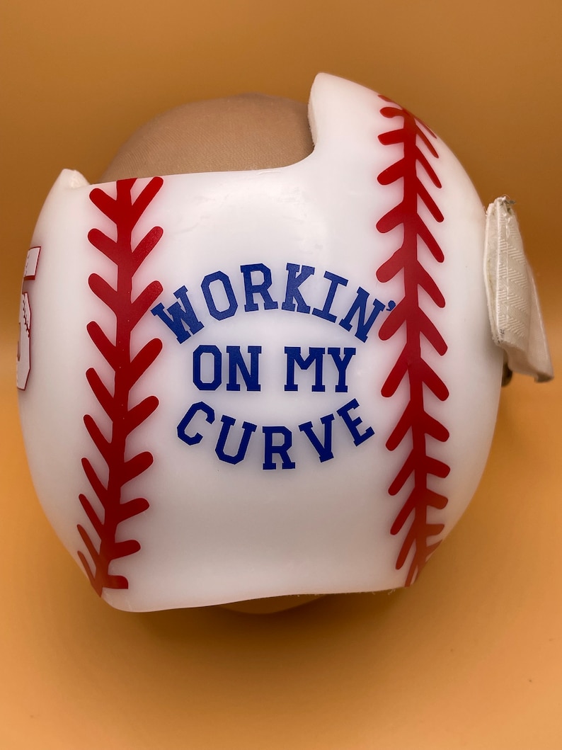 May include: White baseball-themed helmet with red stitching and the text "WORKIN' ON MY CURVE" in blue.