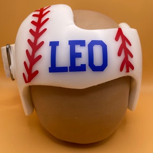 May include: White helmet with a baseball design. The helmet has red stitching and a blue name "LEO" on the front.