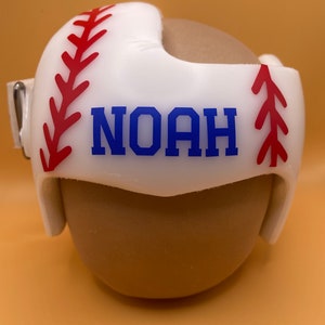 May include: White baseball helmet with red stitching and a blue name 'NOAH' on the front.