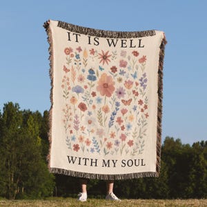 Pode incluir: Cobertor tecido creme com um design floral em várias cores, incluindo rosa, azul e laranja. O cobertor apresenta o texto "IT IS WELL WITH MY SOUL" em preto. As bordas têm franjas.