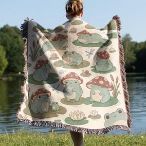 May include: A woven throw blanket featuring a repeating pattern of light green frogs wearing red and white spotted mushroom caps, interspersed with water lilies, cattails, dragonflies, and snails on a cream-colored background with fringed edges.