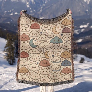 May include: A woven throw blanket featuring a repeating pattern of smiling clouds, crescent moons, and small stars in shades of cream, brown, blue, and tan. The rectangular blanket has a fringed edge along the top and bottom borders.