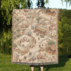 May include: A woven throw blanket featuring an isometric illustration of an ancient village with stone buildings, narrow pathways, olive trees, and livestock. The scene includes figures travelling with camels, market stalls, and a wooden stable structure.