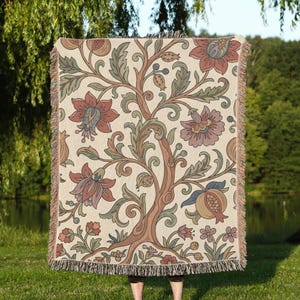 May include: A woven throw blanket featuring a cream-colored background with a repeating botanical pattern of stylized flowers, scrolling vines, and pomegranate-like fruits in muted shades of terracotta, sage green, and dusty blue with fringed edges.