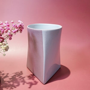 May include: A light gray ceramic vase with a triangular base. The vase is split down the middle, with one side a lighter gray and the other side a darker gray.