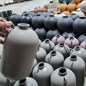 Może przedstawiać: Kolekcja ceramicznych wazonów w różnych rozmiarach i kolorach, w tym szary, jasnofioletowy i czarny. Wazy mają gładkie, matowe wykończenie i są ułożone na wystawie.