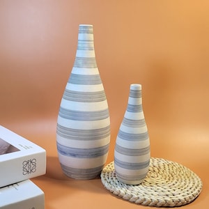 May include: Two white ceramic vases with black stripes. The larger vase is taller and wider than the smaller vase. Both vases are sitting on a woven straw mat.