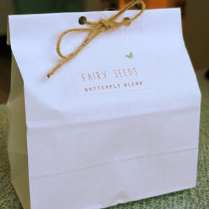 May include: A white paper bag labelled "FAIRY SEEDS BUTTERFLY BLEND" with a green butterfly illustration. The bag is tied with a natural twine bow. The bag is on a green surface.
