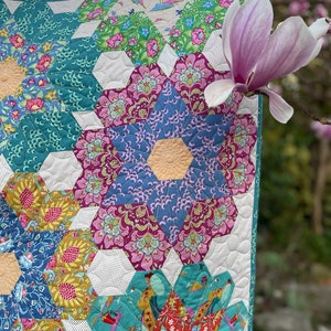 May include: A quilt with a hexagonal pattern featuring a variety of floral prints in shades of blue, green, pink, and yellow. The quilt is quilted with white thread and has a white background. A pink magnolia flower is in the background.