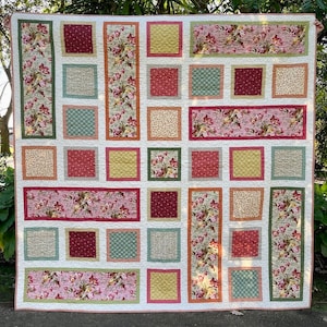 May include: A quilt with a patchwork design featuring various floral patterns in shades of pink, green, and yellow. The quilt is framed with a white border and has a quilted texture.