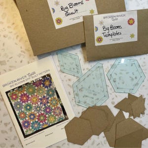 May include: A quilting kit with a pattern, templates, and paper pieces. The pattern cover displays a colourful floral design. The kit includes clear plastic templates and brown paper pieces. The boxes are labelled "Big Blooms Quilt" and "Big Blooms Templates."