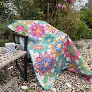 May include: A colourful quilt with a floral pattern is draped over a black chair in a garden setting. The quilt is made of various fabrics in shades of pink, blue, yellow, and green. The chair is sitting on a bed of small stones.