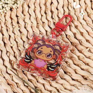 May include: A clear acrylic keychain featuring a cartoon character with glasses, a pink top, and black wings, surrounded by red decorative elements. The keychain has a red clasp and the text "AYEHIKU" is visible.