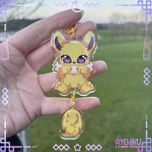 May include: A yellow cartoon character keychain with a fluffy tail and large purple eyes. The keychain is suspended by a yellow chain. The character has a white frill around its neck and is holding its paws up. The text "AYEHIKU" is visible.
