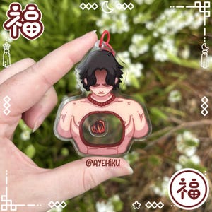 Może przedstawiać: Przezroczysty akrylowy urok przedstawiający stylizowaną ilustrację postaci z czarnymi włosami i czerwonym naszyjnikiem z koralików. Klatka piersiowa postaci ma okrągły wzór z grafiką pięści. Urok ma czerwoną pętlę do zawieszenia. Widoczny jest tekst "@AYEHIKU".