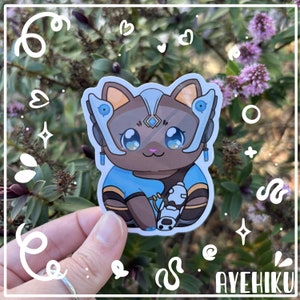 May include: A sticker of a brown and white cat wearing a blue and white spacesuit. The cat has large, round eyes and a small, pink nose. The sticker is on a green and brown background with white hearts and stars.