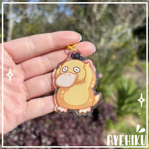 May include: A yellow Psyduck acrylic charm with a yellow keyring. The charm features a cartoon illustration of Psyduck with a large bill and surprised expression. The charm is held in a hand against a blurred green background.