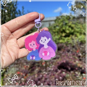 May include: A clear acrylic keychain featuring two cartoon figures, one with pink hair and the other with purple hair, both holding mugs. The keychain has a purple clasp and the words "HIKU ARTISTRY" are printed at the bottom.