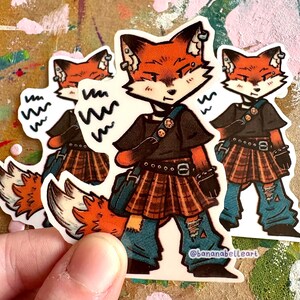 May include: A die-cut sticker featuring an anthropomorphic fox character with orange fur, wearing a black t-shirt, a brown plaid skirt, and blue distressed pants. The character has a blue shoulder bag and the text @bananabelleart is printed at the bottom.