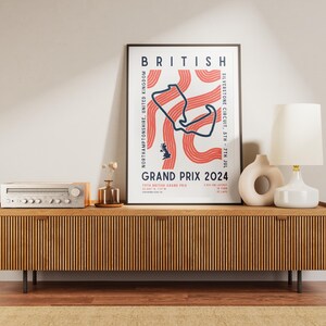 May include: A poster with a minimalist design featuring a red and white track map for the British Grand Prix 2024. The poster is framed in black and is displayed on a wooden shelf with a vintage stereo and a lamp.