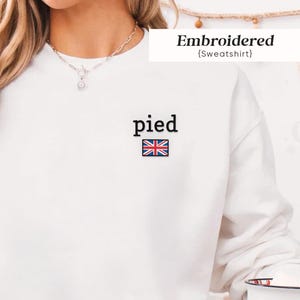 May include: A white long-sleeve sweatshirt featuring the word pied in black text above a small embroidered Union Jack flag on the left chest. The garment is paired with light blue denim jeans and a silver chain necklace.