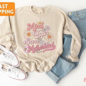 May include: A beige sweatshirt with the text "Mind Your Own Motherhood" in pink and white lettering with a floral design.