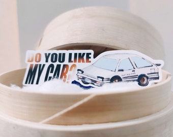 Op de afbeelding: Een witte auto sticker met de tekst "Do you like my car?" en een tekening van een witte auto met zwarte accenten. De sticker zit op een lichtbruin houten oppervlak.