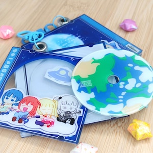 May include: A collection of anime-themed CD cases and a CD. The cases are blue with cartoon illustrations of a band and a spaceship. The CD has a planet design with green continents and blue oceans. A blue lanyard is attached.