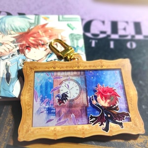 May include: A gold-colored keychain with a wooden frame featuring a colorful illustration of a clock tower, a flying figure with red hair, and purple feathers. The keychain has a metal clasp.