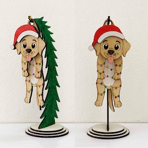 May include: Wooden Christmas ornament featuring a cartoon dog wearing a Santa hat and wrapped in colorful string lights. The dog is light brown with a black nose and tongue sticking out. The ornament is on a round, tiered base.