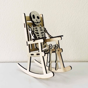 Könnte beinhalten: Eine Holzfigur eines Skeletts sitzt in einem Schaukelstuhl aus Holz. Das Skelett hat einen detaillierten Schädel und Brustkorb. Der Stuhl ist aus hellem Holz mit einer Lamellenlehne und geschwungenen Schaukeln gefertigt, ideal für Halloween.