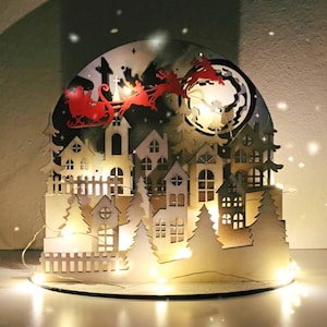 May include: Wooden Christmas village scene with Santa in a sleigh pulled by reindeer, cut from wood. The village features houses, trees, and a fence, illuminated by warm white lights. A full moon is in the background.