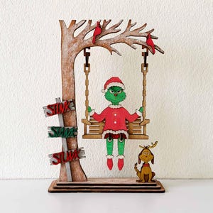 Puede incluir: Decoración navideña de madera con un personaje verde con traje de Papá Noel en un columpio, con un perro y un árbol. El árbol tiene letreros que dicen "STINK, STARE, STUNK". Dos pájaros rojos se posan en las ramas.