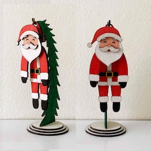 Può includere: Due statuette di Babbo Natale in legno. Ogni Babbo Natale indossa un abito rosso con cintura e stivali neri, barba bianca e cappello rosso. Uno tiene un albero di Natale verde. Le statuette sono su una base rotonda a più livelli.