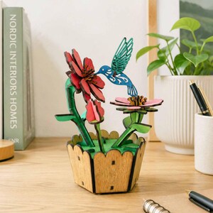 May include: A wooden hummingbird and flower sculpture. The blue and green hummingbird is positioned near a pink flower, with red flowers and green stems in a wooden planter. The sculpture is on a desk.