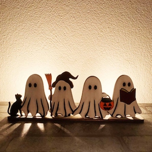 Op de afbeelding: Een set van vijf witte houten spookfiguren met zwarte ogen. De spoken zijn in een rij op een houten basis gerangschikt. Het spook links houdt een zwarte kat vast. Het volgende spook houdt een bezem vast. Het derde spook draagt een heksenhoed. Het vierde spook houdt een kleine oranje pompoen vast. Het laatste spook houdt een boek vast.