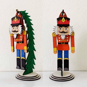 Peut inclure: Figurine de casse-noisette de Noël en bois rouge, noir, jaune et bleu. La figurine a une barbe et une moustache blanches et un chapeau rouge. Le casse-noisette est posé sur une base en bois ronde. Un sapin de Noël en bois vert est fixé au dos de l'un des casse-noisettes.