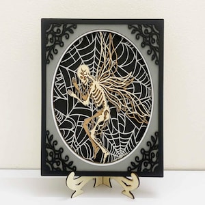 Puede incluir: Una obra de arte de madera en capas que presenta un hada esqueleto dentro de un diseño de telaraña. La obra de arte tiene un marco negro con esquinas decorativas, un fondo gris y un soporte de madera. El hada tiene alas detalladas y una estructura esquelética.