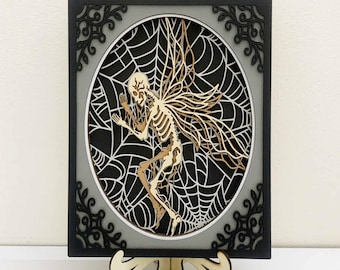May include: A layered wooden art piece featuring a skeleton fairy within a spiderweb design. The artwork has a black frame with decorative corners, a gray background, and a wooden stand. The fairy has detailed wings and a skeletal structure.