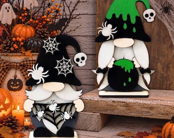 May include: Two wooden gnome decorations for Halloween. One gnome wears a black hat with white spiderwebs and spiders, holding a spiderweb pattern. The other gnome wears a green and black hat with a white spider, holding a green bubbling cauldron.