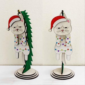 Könnte beinhalten: Zwei Holzkatzen-Ornamente mit Weihnachtsmützen und Lichterketten. Die Katzen haben mürrische Ausdrücke und hängen an einem Ständer. Eine Katze steht neben einem grünen Weihnachtsbaum. Die Ornamente sind etwa 15 cm hoch.