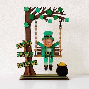 Könnte beinhalten: Holzdekoration zum St. Patrick's Day mit einem Kobold auf einer Schaukel, einem Baum mit Kleeblättern und einem Goldtopf. Das Schild lautet "Lucky Irish St. Patrick". Der Kobold trägt einen grünen Anzug und Hut.
