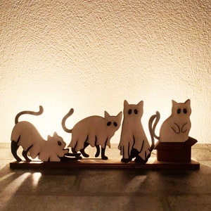 Könnte beinhalten: Ein Set aus vier Holzkatzenfiguren, von denen zwei Geister sind, die auf einer Holzbasis stehen. Die Katzen sind in Braun- und Weißtönen bemalt.