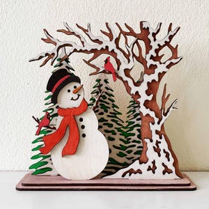 Peut inclure: Scène hivernale en bois représentant un bonhomme de neige avec un chapeau noir et une écharpe rouge, devant des sapins et un arbre enneigé. Deux cardinaux rouges sont perchés sur des branches. La base est de couleur marron clair.