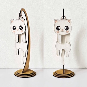 May include: Wooden cat figurine in a cartoon style. The white cat has large black eyes, pink cheeks, and a tail. The cat is hanging from a wooden stand with a circular base. The figurine is a decorative item.