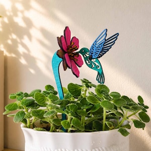 Puede incluir: Un tutor decorativo para plantas con un colibrí de madera cortado con láser en tonos azul y verde azulado, cerca de una flor rosa vibrante con un centro oscuro. El tutor está insertado en una planta verde en una maceta blanca.