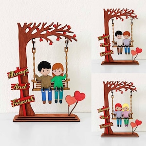 May include: Wooden decorative piece featuring a couple on a swing under a tree with heart-shaped leaves. The words "Always And Forever" are displayed on a sign. Includes heart-shaped balloons. The couple is wearing blue pants and different colored tops.