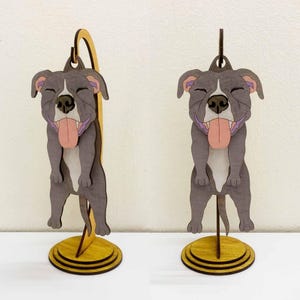 May include: Wooden dog figurine with a smiling face and tongue sticking out. The dog is gray with pink and purple accents, mounted on a yellow wooden base. The figurine is designed to stand upright, showcasing a playful and cheerful aesthetic.