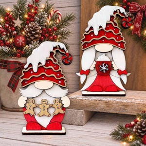 Può includere: Due decorazioni in legno a forma di gnomo nei colori rosso e bianco. Uno gnomo tiene in mano una catena di tre omini di pan di zenzero, mentre l'altro regge una tazza rossa con un fiocco di neve bianco. Entrambi indossano alti cappelli.