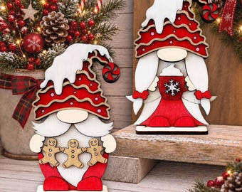 May include: Two wooden gnome ornaments featuring red and white color schemes. One gnome holds a string of three gingerbread men, while the other holds a red mug with a white snowflake design. Both wear tall hats with white snow-like trim.